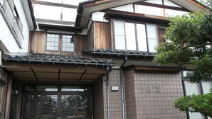 Ein traditionelles japanisches Haus mit einem gefliesten Eingangsbereich, Holzbalken und einem Baum vor der Fassade.