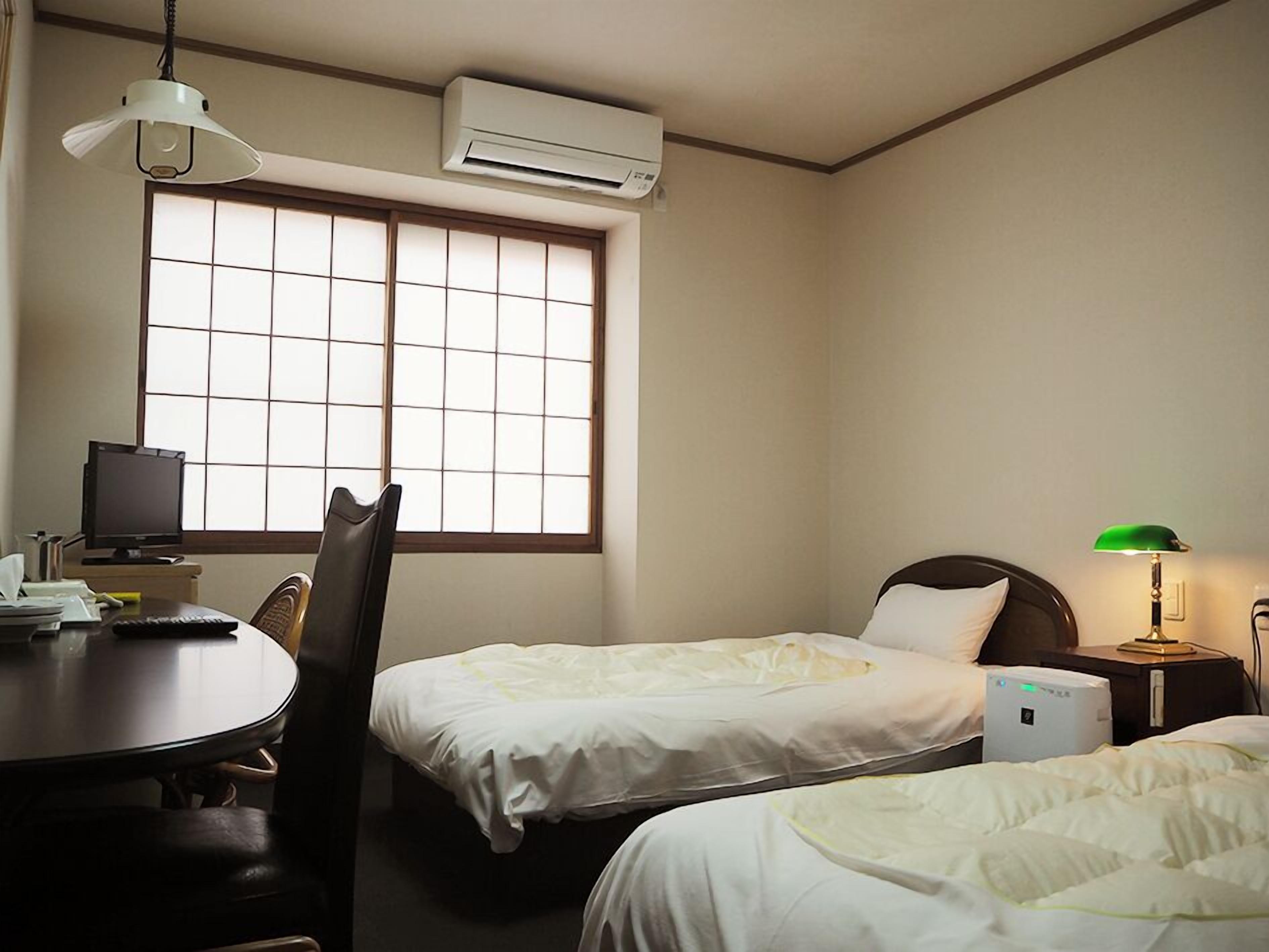 A hotel room with two beds, a desk with a computer, a chair, an air conditioner, and a window with blinds.