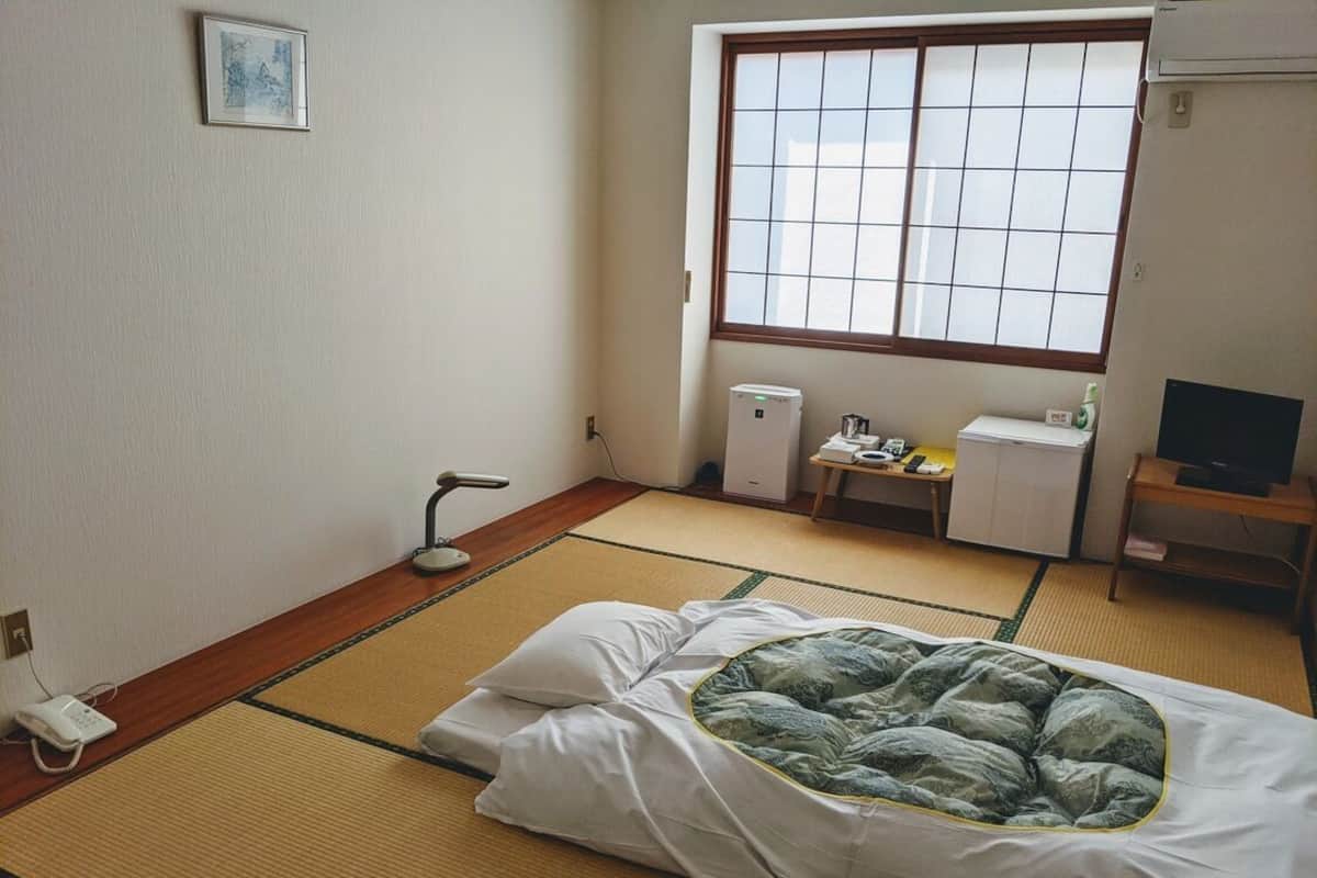 A traditional Japanese-style room with tatami flooring, a futon bed, a small desk with a computer, and a window with a grid pattern.
