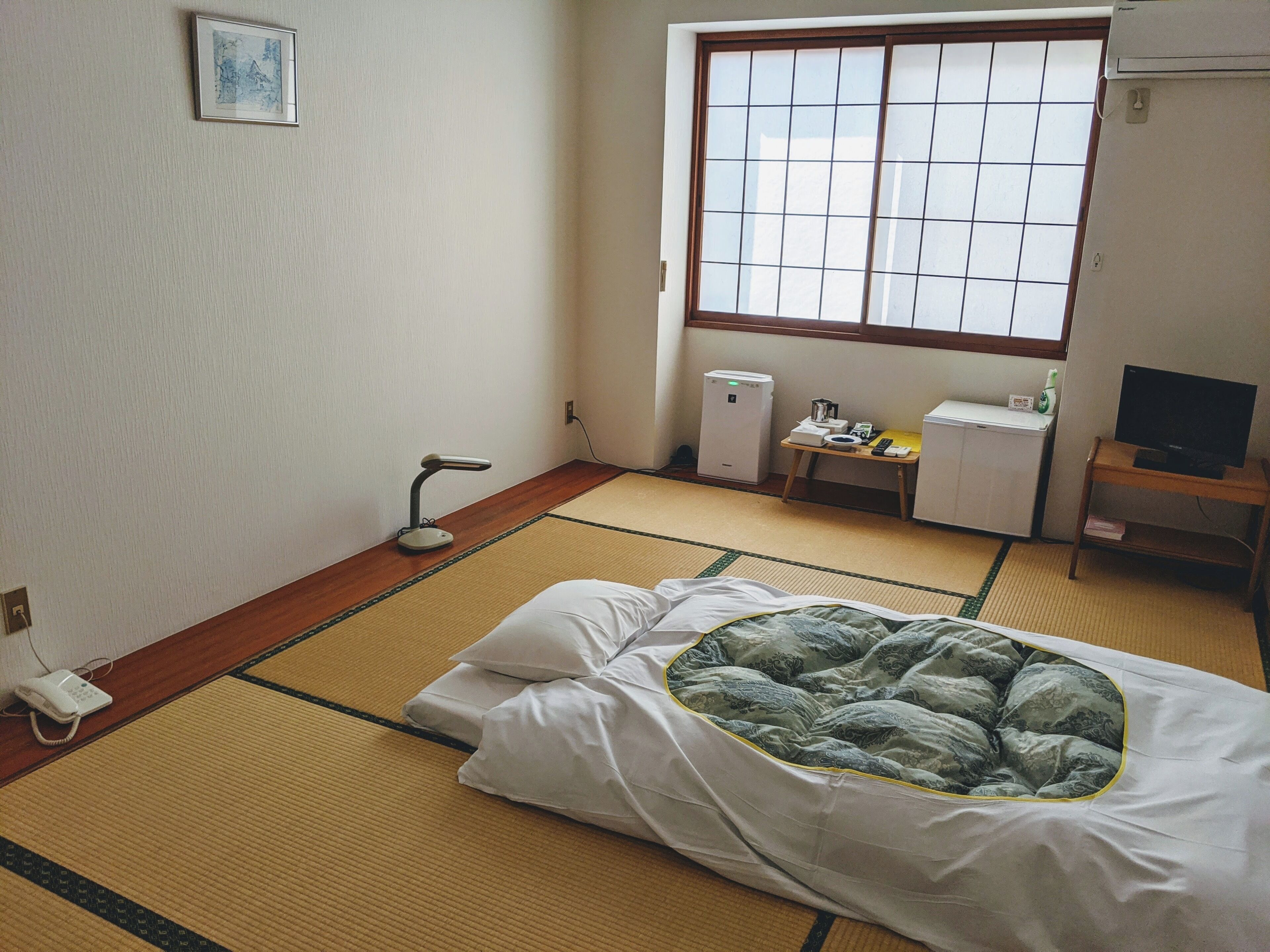 Una habitación con suelo de tatami, una cama con manta verde estampada, un escritorio con computadora, un pequeño calentador y una ventana con vistas.