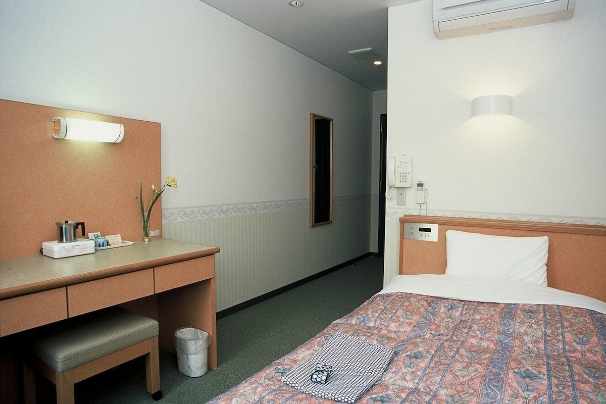 A hotel room with a bed, bedside table, desk, and a trash bin.