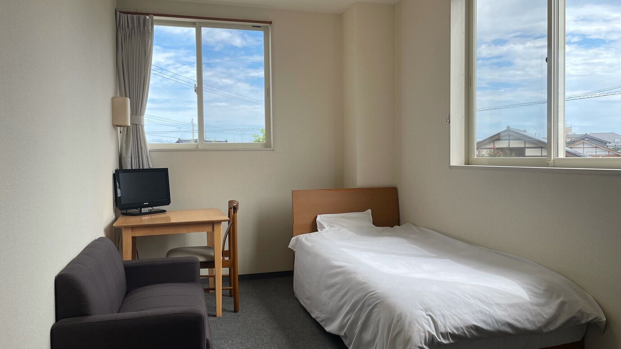 A hotel room with a single bed, a desk with a television, a chair, and a window with a view of buildings.