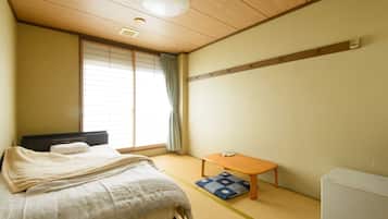 A room with a bed, a small table, a futon, a futon blanket, a futon pillow, a futon mat, a window with blinds, and a wall-mounted shelf.