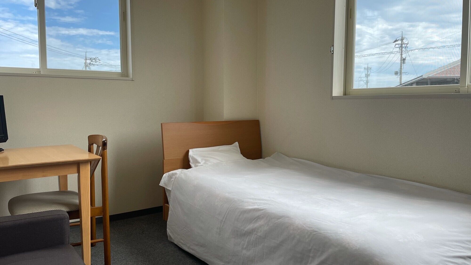 Una cama individual con ropa de cama blanca, un escritorio de madera con una silla, y una ventana con vista a líneas eléctricas y un edificio.