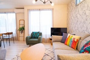 A living room with a sofa, armchair, and a TV. There is a window with curtains, a plant, and a ladder against the wall. - Golden Drop S (Kin)