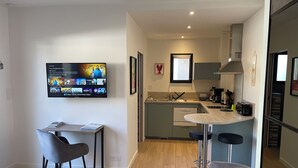 A modern kitchen with a built-in oven, a dining area with a table and chairs, and a flat-screen TV mounted on the wall displaying a streaming service interface. - Quiet studio in the heart of Arradon (Arradon)