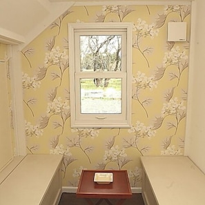 Una habitación con papel tapiz floral, una ventana con vista a árboles, una pequeña mesa de madera y un banco empotrado.