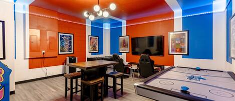 A room with a pool table, a foosball table, and a television. There are chairs and a table with a few people around it.