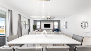 Un salon moderne avec une grande table blanche, des fauteuils gris, un ventilateur de plafond et un téléviseur fixé au mur.