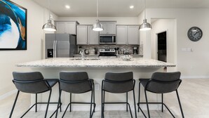 Una cocina moderna con una isla grande, taburetes de bar, iluminación colgante y un reloj en la pared.