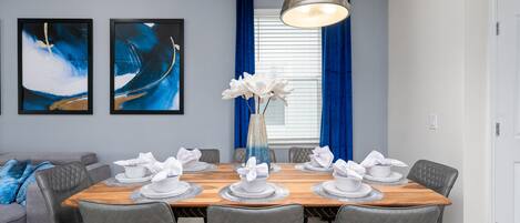 Un comedor con una mesa de madera dispuesta para seis, sillas tapizadas de gris, un florero con flores blancas y pinturas abstractas en la pared.