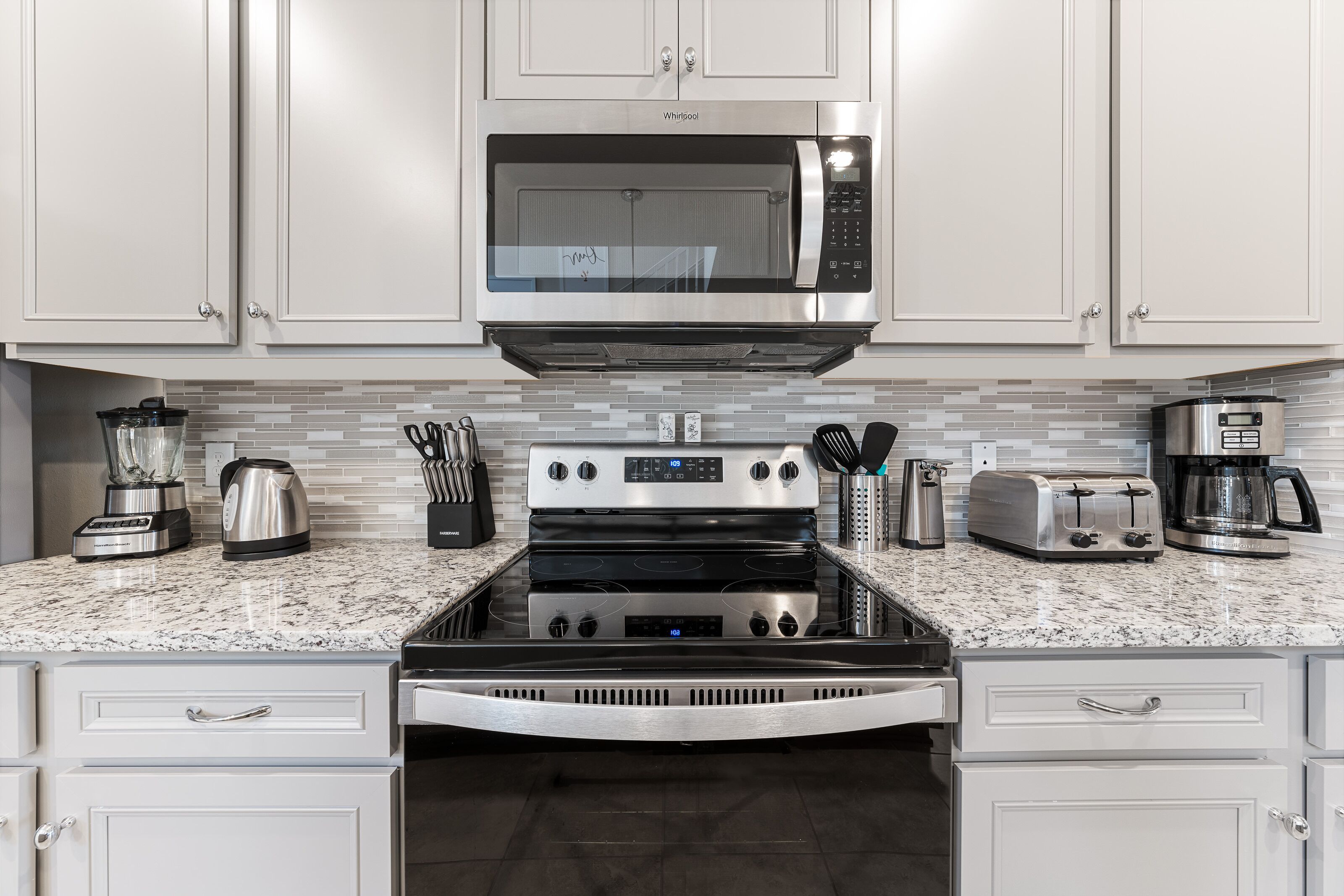 A modern kitchen with white cabinets, a stainless steel microwave, a built-in oven, a coffee maker, and a toaster.