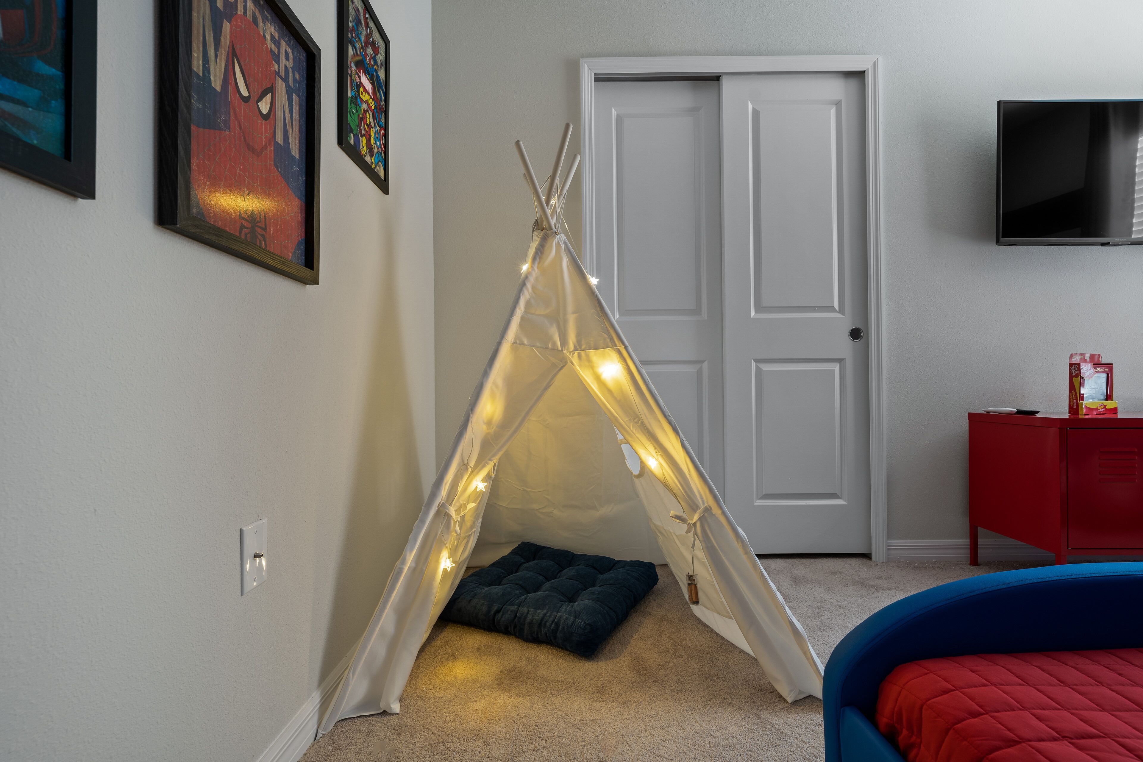 Une pièce comprenant une tente de type tipi, un lit, une commode rouge, un téléviseur et des tableaux encadrés accrochés au mur.
