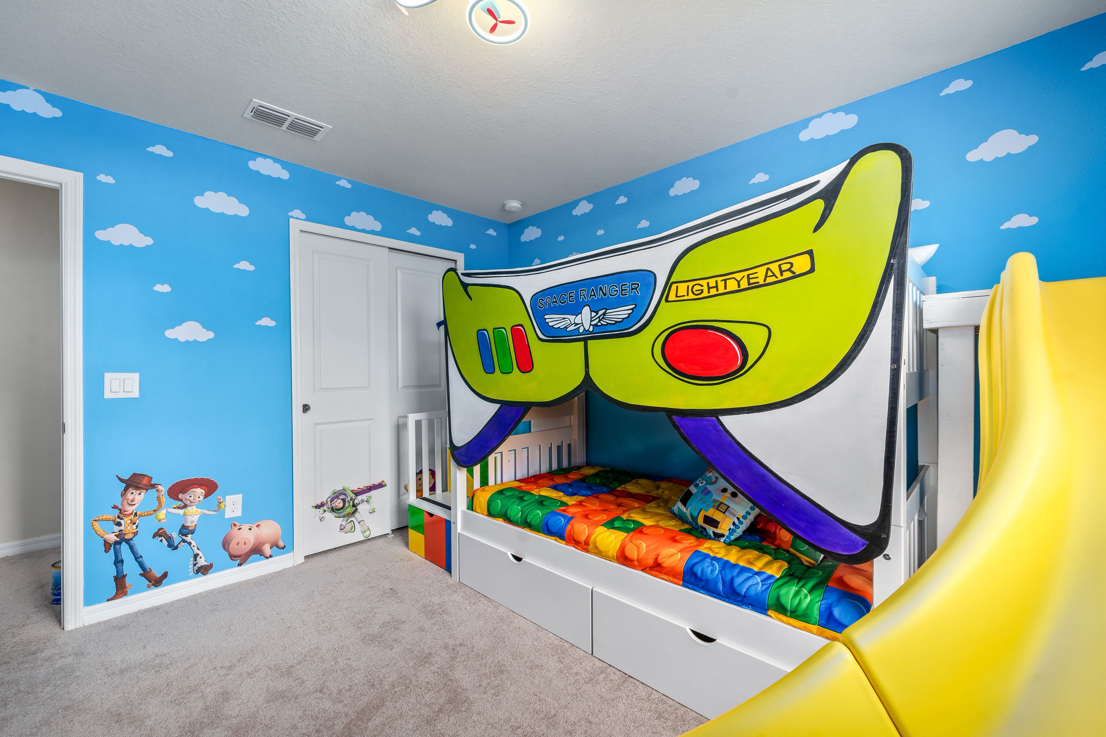 Habitación infantil con cama decorada con motivos espaciales, una puerta blanca y un muro con un vinilo de cielo azul y nubes.