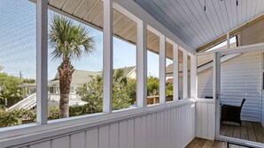 A covered porch with white railings and a wooden floor, overlooking a residential area with houses and palm trees. - Steps to the Beach | King Beds | Pool Access (Tybee Island)