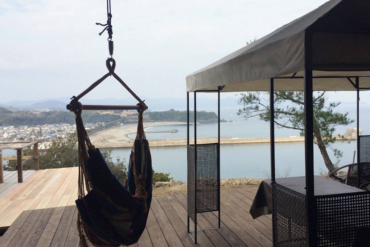 A hammock hanging on a wooden deck overlooking a coastal view with a covered seating area.