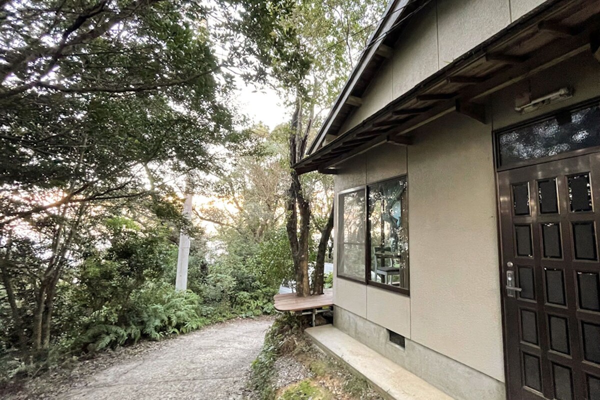 A building with a large window and a door, surrounded by trees and a gravel path.