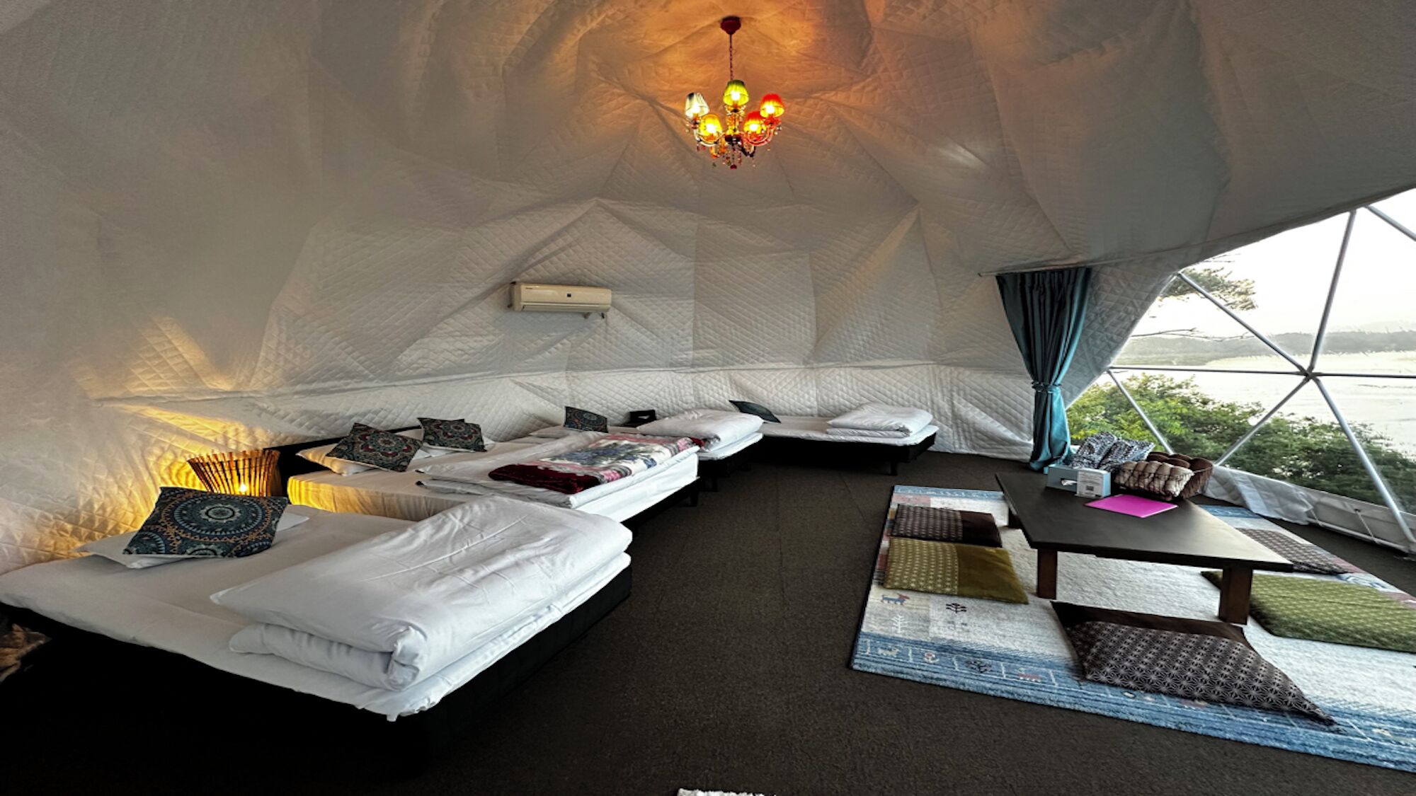 Ein Zimmer mit vier Betten, einem Tisch, einer Chandelier und Blick durch ein großes Fenster.