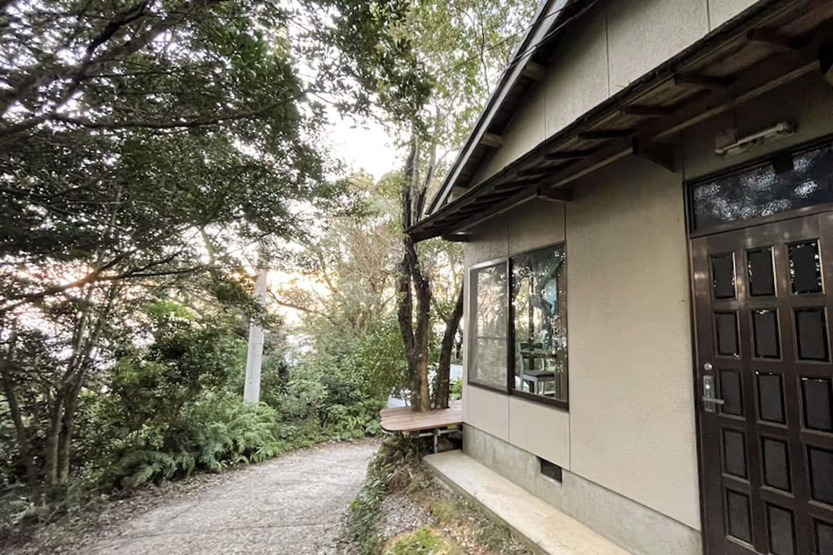 A building with a large window and a door, surrounded by trees and a gravel path.