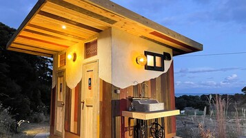 Un pequeño edificio rústico con techo inclinado, una puerta y un aplique sobre ella, y una ventana con un aplique.