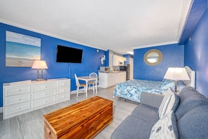 A room with a bed, a sofa, a wooden chest, a desk with chairs, a television, a kitchen area, and a blue wall with a beach painting.