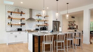 A modern kitchen with white cabinetry, a large island with bar stools, hanging pendant lights, and a brick backsplash.