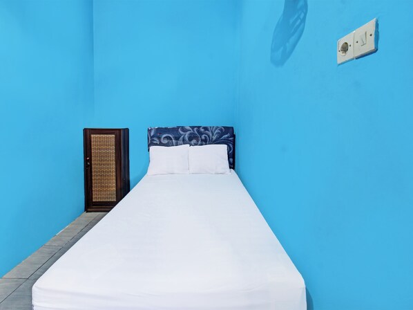 Una cama individual con colchón y almohadas blancas, una puerta de madera con panel tejido y un enchufe de pared.