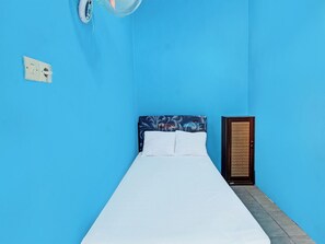 A single bed with a white mattress and a headboard with a floral pattern, a small bedside table, and a wall-mounted fan. - Spot On 91811 Rumah Inap Bunda Nurhayati (Tanak Awu)