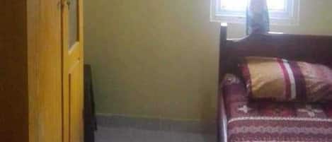 A bedroom with a wooden wardrobe, a bed with patterned bedding, a window with blue curtains, and an air conditioning unit.