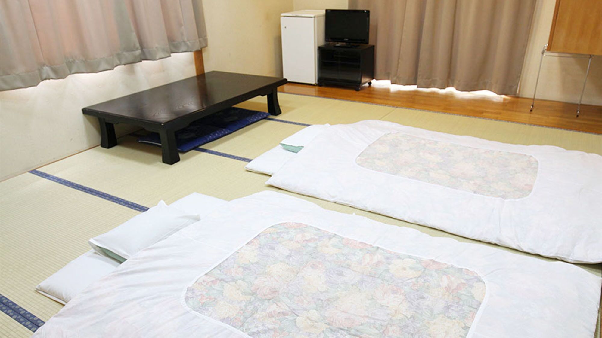 A room with tatami mats, a low table, and two futons with floral patterns.