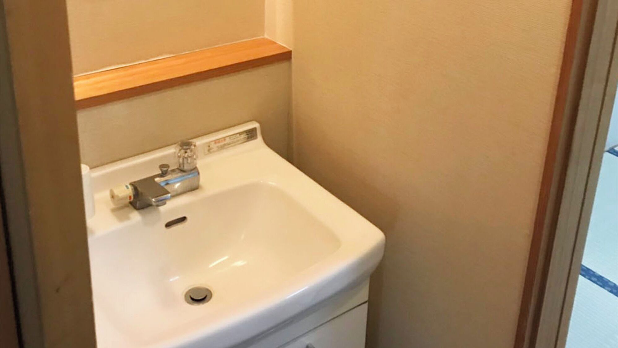 Une salle de bain avec un lavabo blanc, une étagère en bois au-dessus et des murs de couleur beige.