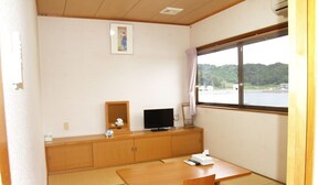 A room with tatami flooring, a wooden table, a TV on a cabinet, and a window with a view of a body of water and hills. - Guesthouse Amy Monbos (Higashikagawa)