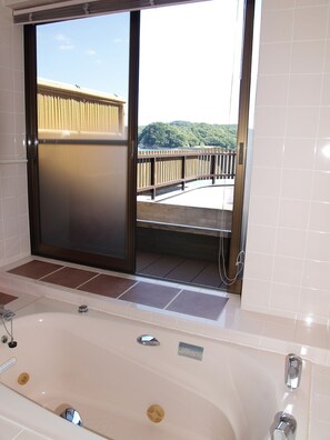 A bathroom with a bathtub, a sliding glass door leading to a balcony with a wooden railing, and a view of a forested hill.