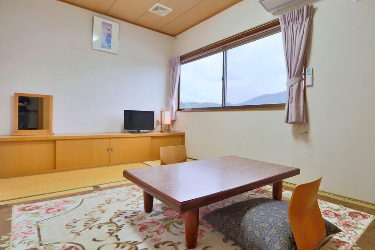 A room with tatami flooring, a low table, a chair, a television, and a window with curtains.