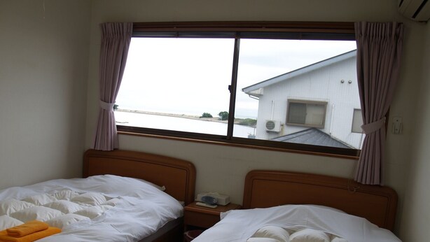 A hotel room with two beds, a window with curtains, and a view of a building across the water. - Guesthouse Amy Monbos (Higashikagawa)
