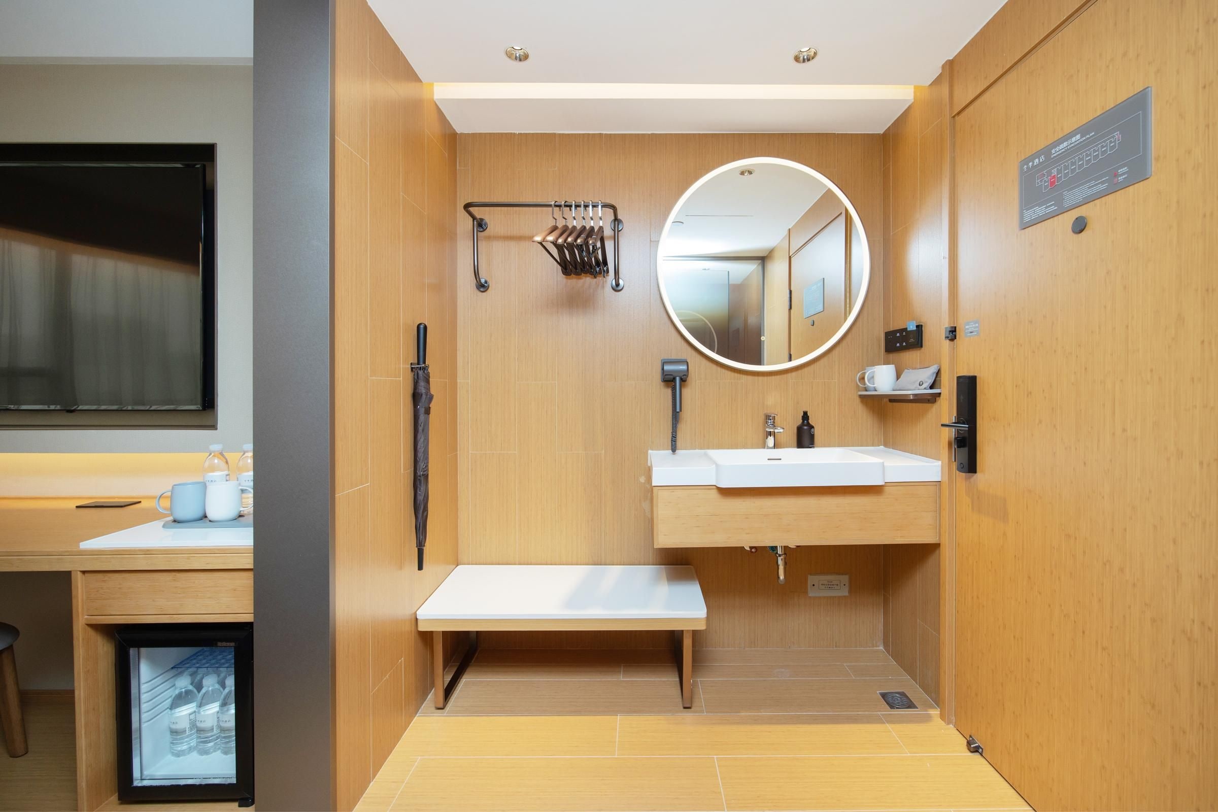 A modern bathroom with a wooden vanity, a round mirror, a towel rack, and a bench.