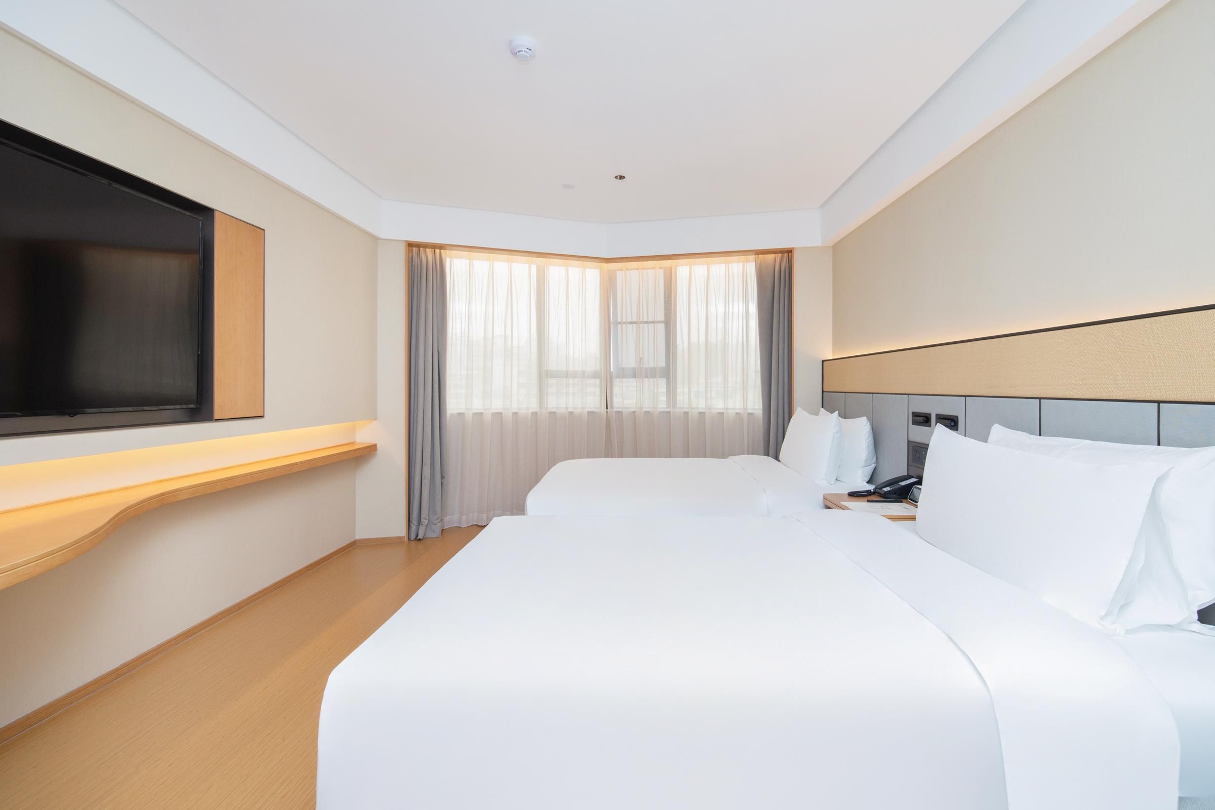 A hotel room with a large bed, a flat-screen TV mounted on the wall, a bedside table with a phone, and a window with sheer curtains.