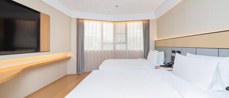 A hotel room with a large bed, a flat-screen TV mounted on the wall, a bedside table with a phone, and a window with sheer curtains.