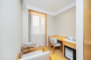Una habitación compacta con un escritorio de madera, una silla, una mesita redonda con una silla blanca, una ventana con cortinas traslúcidas y un mini refrigerador empotrado.