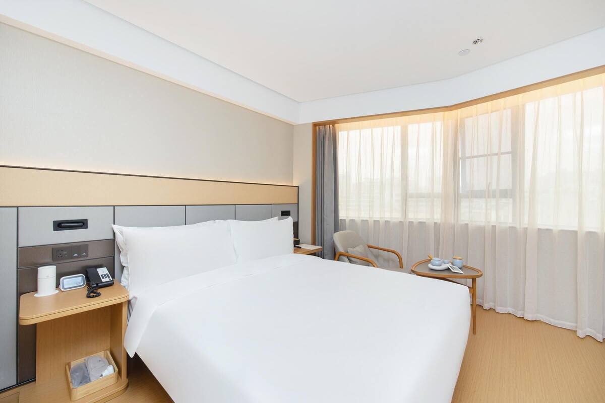 A hotel room with a large bed, bedside table with a phone and a coffee maker, a chair, a small table with a cup and saucer, and a window with sheer curtains.