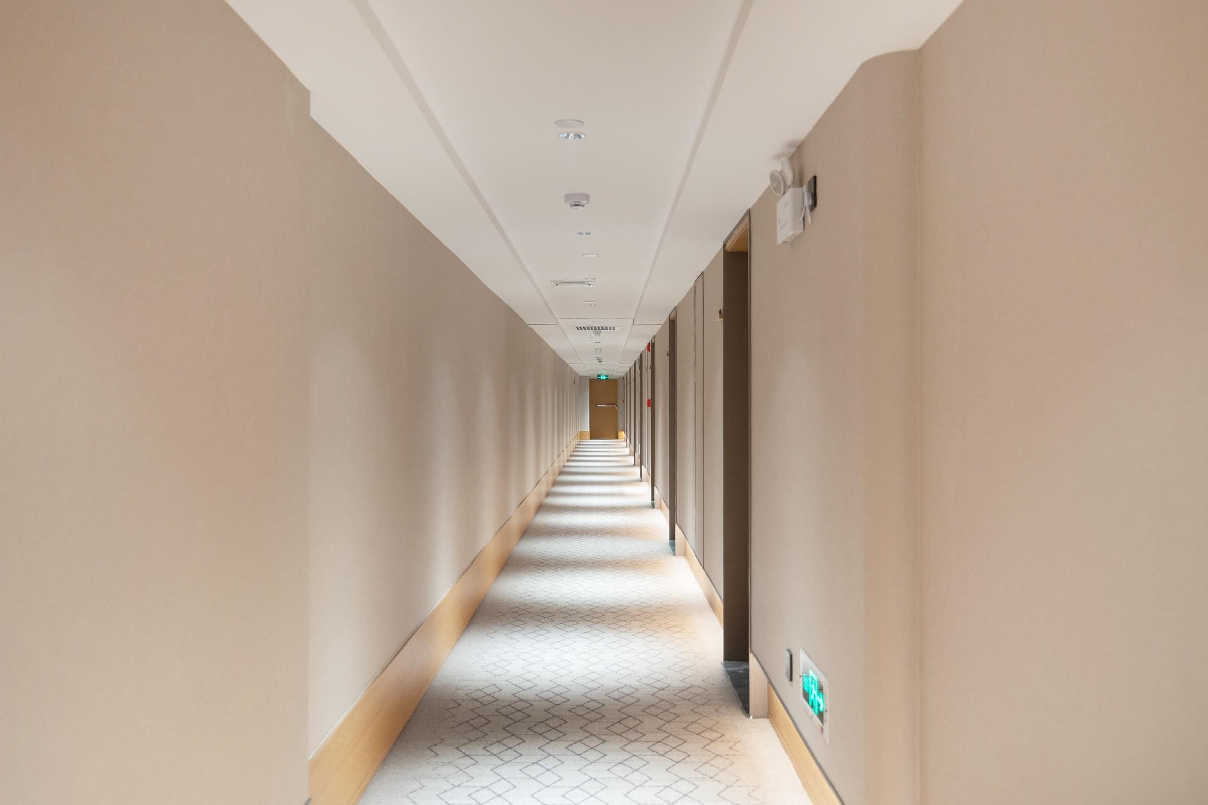 Un largo y estrecho pasillo de hotel con piso estampado, paredes lisas e iluminación empotrada en el techo.