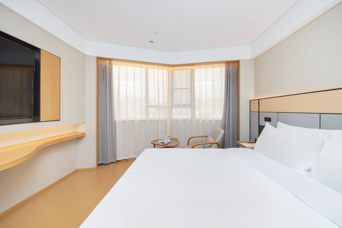 A hotel room with a large bed, a TV mounted on the wall, a small table with a lamp, and a chair.