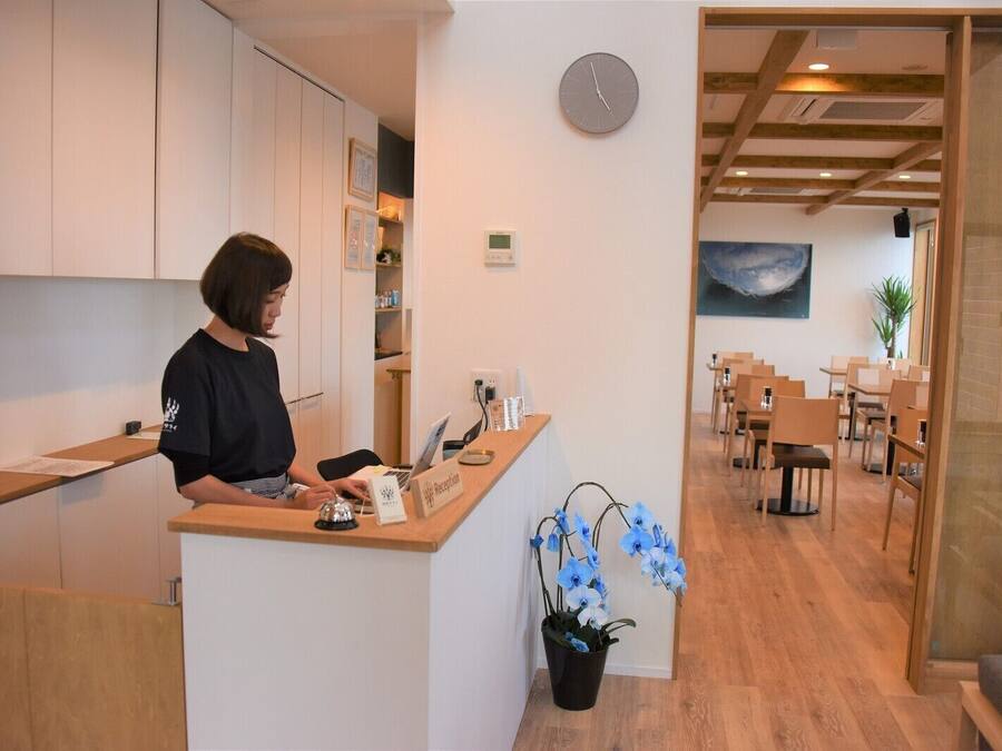 A reception desk with a clock, a potted plant, and a view of a dining area with tables and chairs.