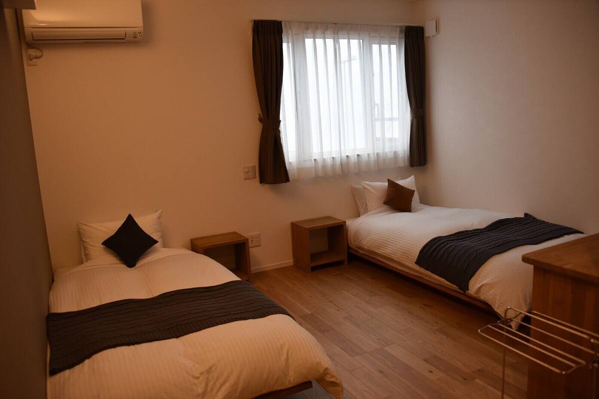 Una habitación con dos camas, una ventana con cortinas, un aire acondicionado de pared y piso de madera.