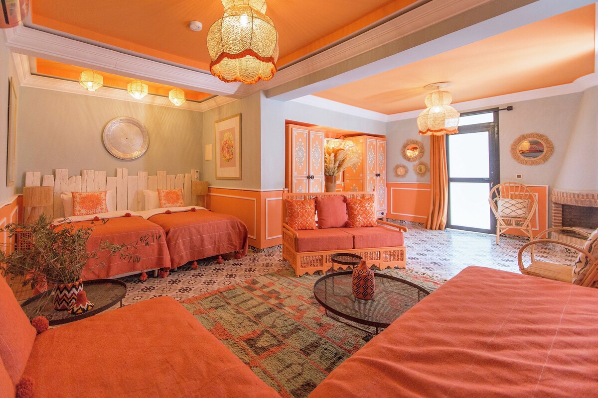 A room with a bed, a sofa, a small table, and a chair. There are decorative items on the walls and a window with curtains.