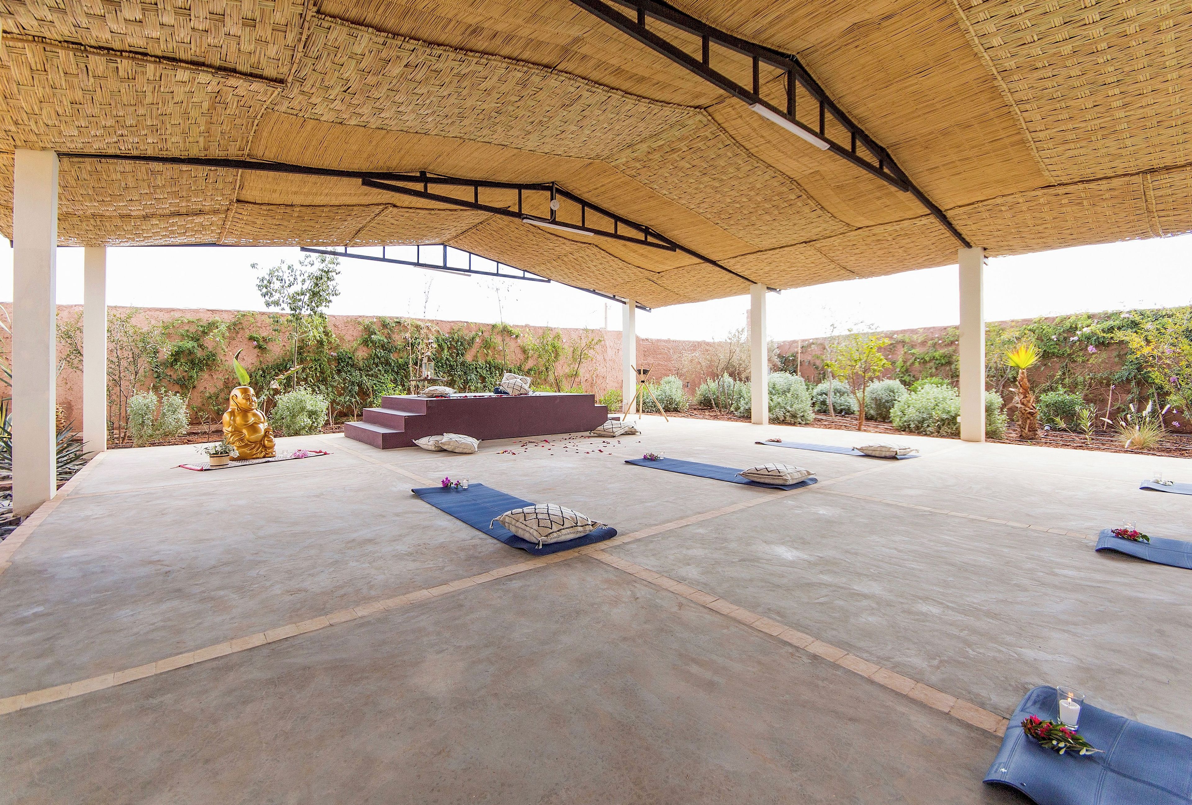 An open-air pavilion with a thatched roof, a Buddha statue, and a meditation area with mats and cushions.