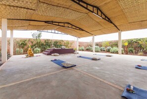 An open-air pavilion with a thatched roof, a Buddha statue, and a meditation area with mats and cushions.