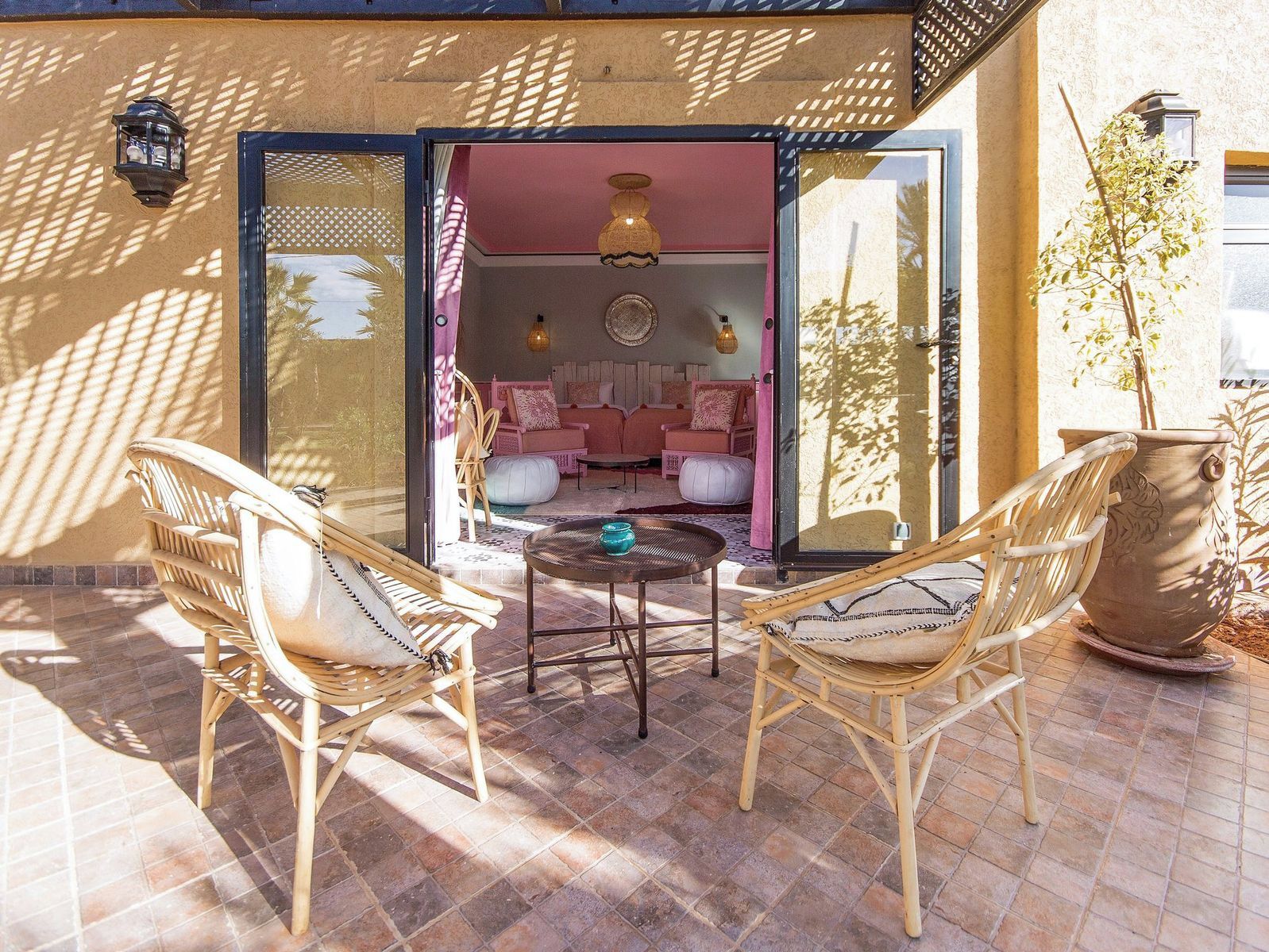 An outdoor seating area with wicker chairs and a round table, a potted plant, and a view into a room with pink walls and a sofa.