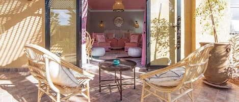 An outdoor seating area with wicker chairs and a round table, a potted plant, and a view into a room with pink walls and a sofa.