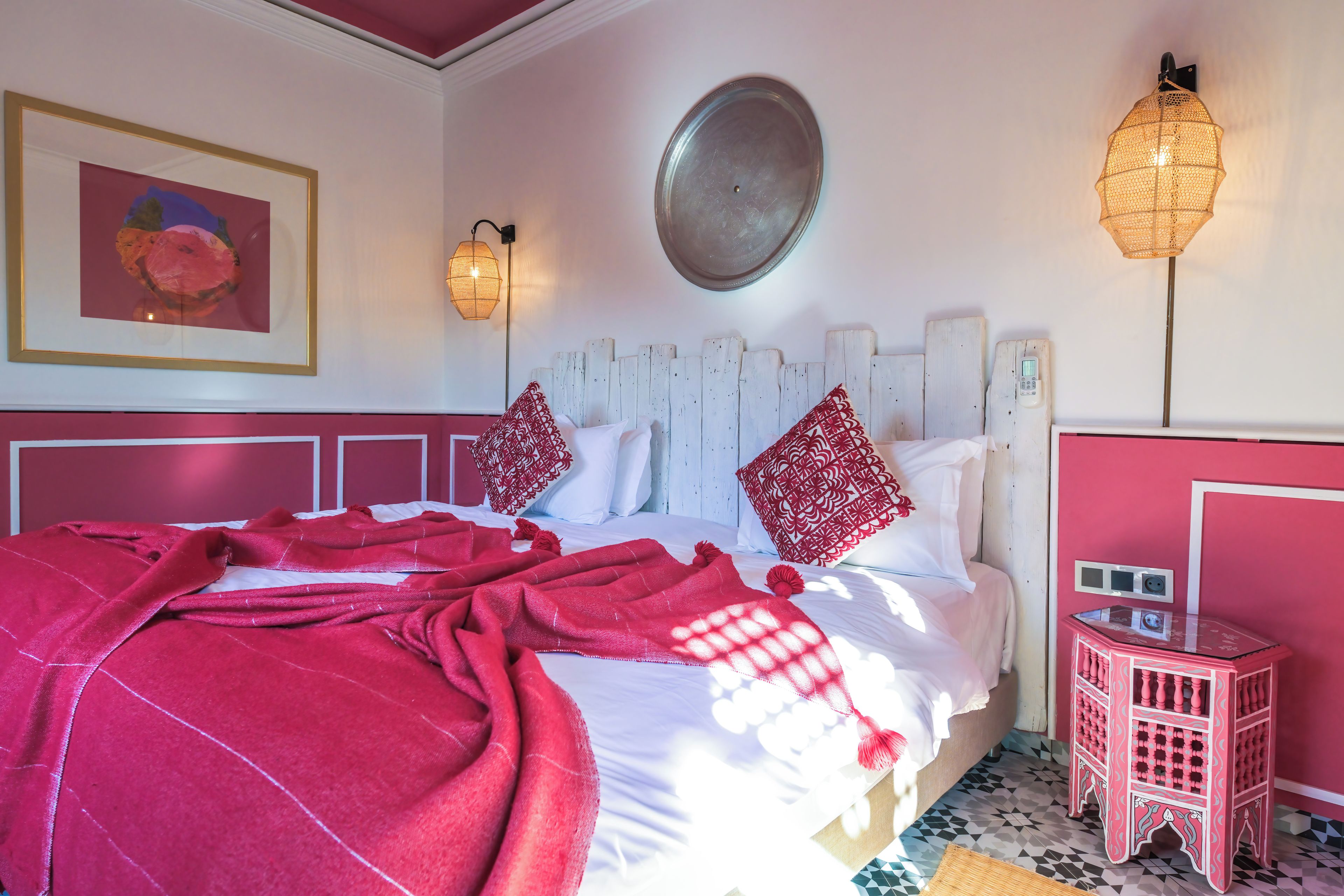 A bed with a red blanket, white pillows, and a wooden headboard. There is a small red nightstand with a lamp and a wall-mounted light fixture.